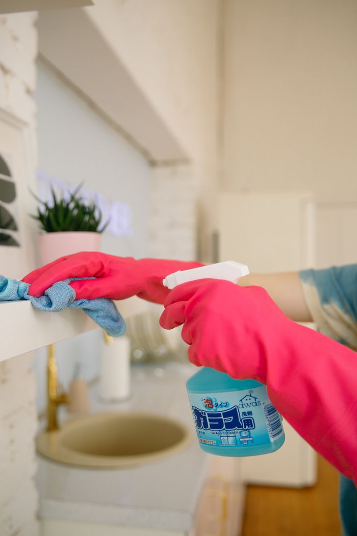 creative-01 Person using spray bottle and cloth for cleaning a kitchen shelf, ensuring hygiene and cleanliness.