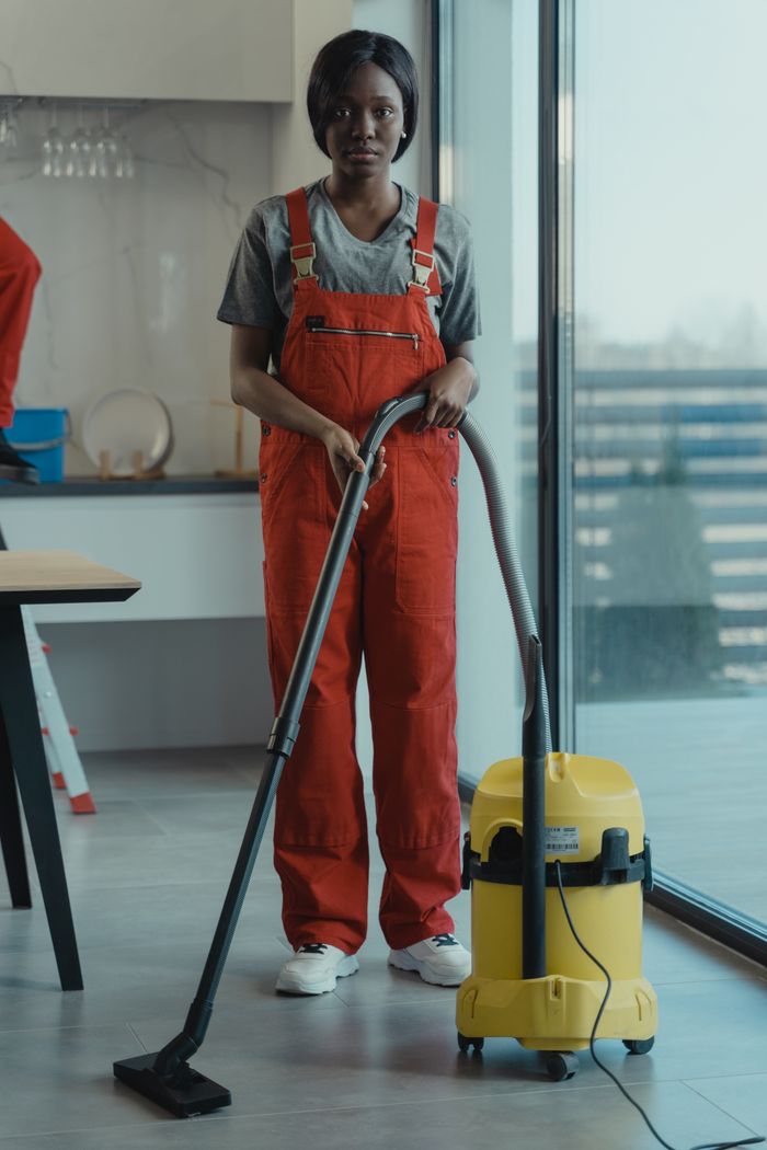 creative-03 An adult woman in red coveralls operates a vacuum cleaner in a modern indoor setting, focusing on cleaning tasks.